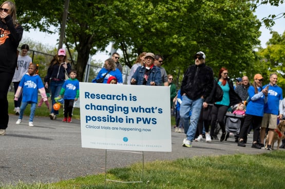 People participating in the One Small Step Maryland walk pass a sign reading “Research is changing what’s possible in PWS” during an outdoor community fundraising and awareness event for Prader-Willi syndrome research.