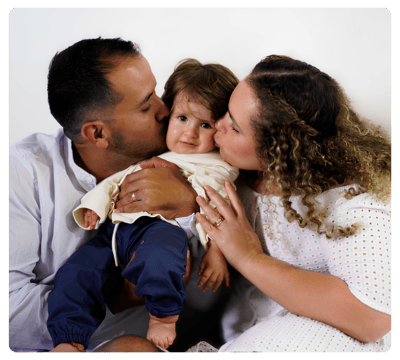 Parents kissing their smiling young child on the cheeks while holding them together.