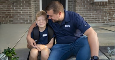 Evan smiles while sitting on a porch step with his backpack as Blake wraps an arm around him and leans in close.