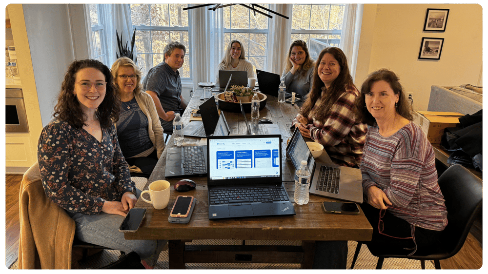 Seven FPWR researchers sit around a table with laptops, phones, and notebooks during a collaborative team meeting in a bright room.
