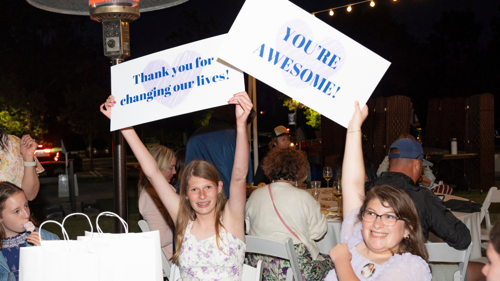 Two young women at the Pars for Peyton golf fundraiser hold signs reading “Thank you for changing our lives!” and “You’re awesome!” during an outdoor evening celebration.