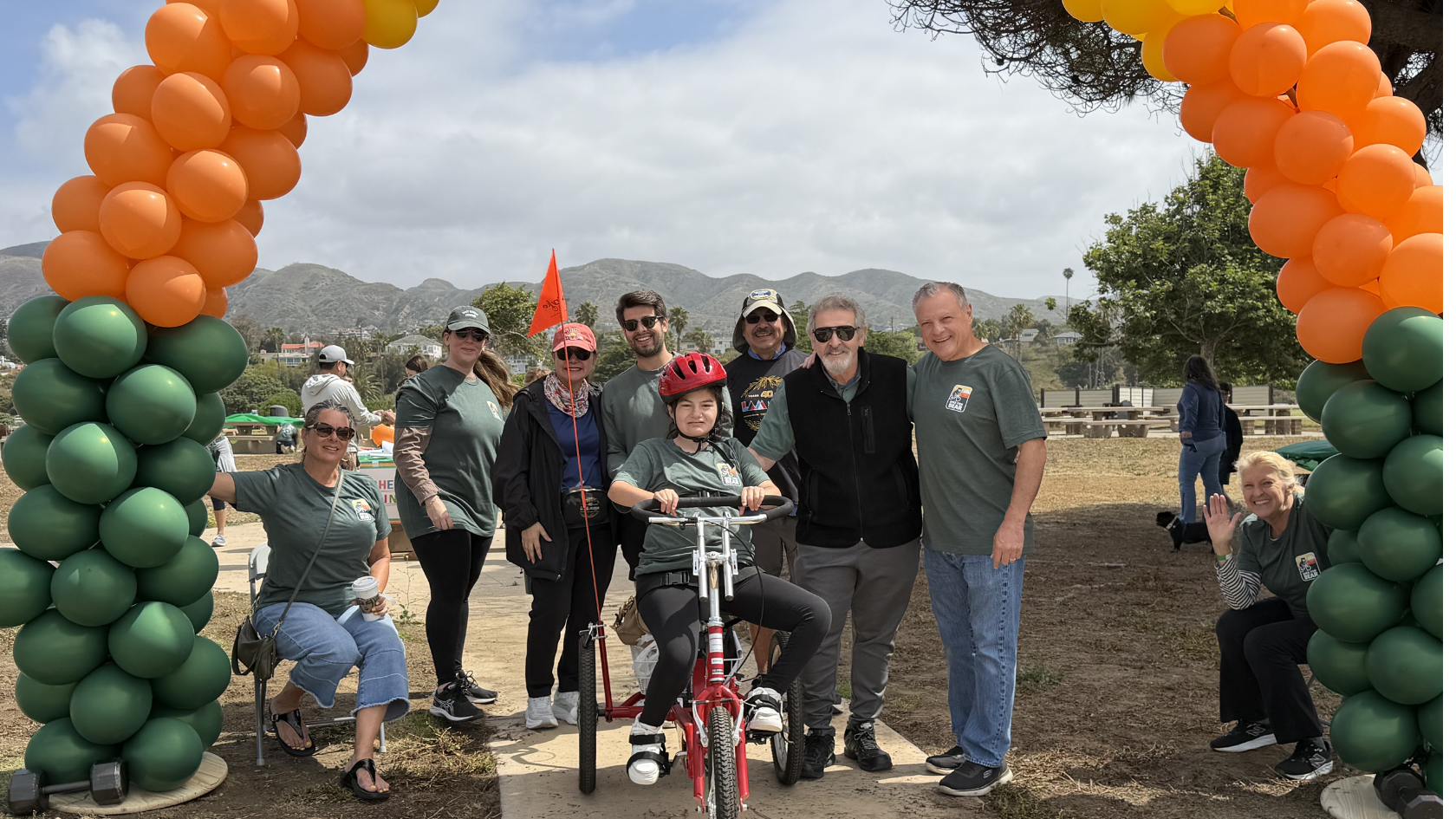 Participants pose beneath an orange-and-green balloon arch at Bike for Bear, with one rider seated on an adaptive tricycle.