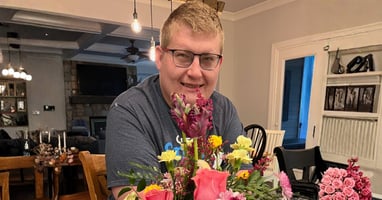 Kyle smiles at the camera while seated at a table with a bouquet of flowers in front of him.