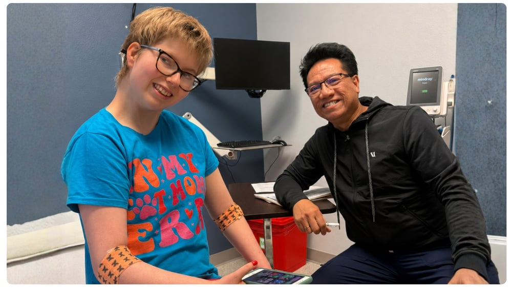 Smiling young woman with glasses sits beside her doctor in a medical exam room, with bandages visible on both arms.
