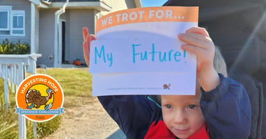 Child holding a sign that reads “We Trot For… My Future!” outdoors, with the Harvesting Hope Distance Challenge logo displayed beside them.