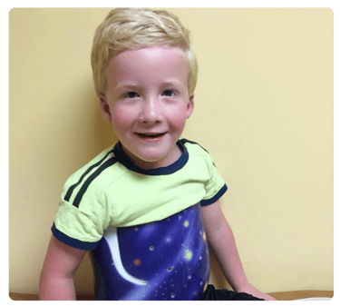 Young blond boy smiles at the camera while seated in a scoliosis brace.