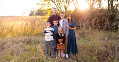 A smiling family of six poses in a grassy field at sunset. The parents stand behind their four children, who range from young child to teenager. Golden sunlight streams through the trees, illuminating the scene with warm, natural light. The family stands close together, creating a welcoming portrait that captures connection, warmth, and togetherness in an outdoor setting.