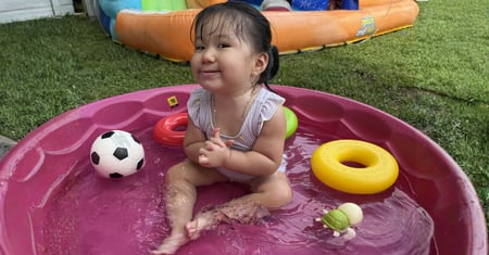Young child smiling while sitting in a small backyard splash pool with colorful floating toys
