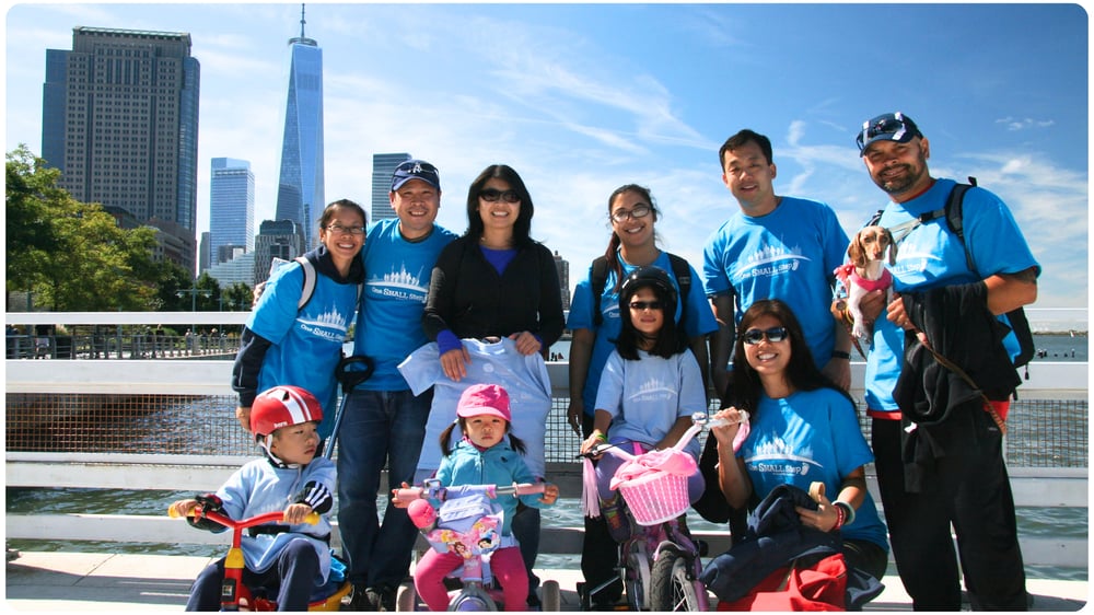 Group of adults and children wearing matching blue fundraising shirts pose together outdoors with bicycles and a city skyline in the background.