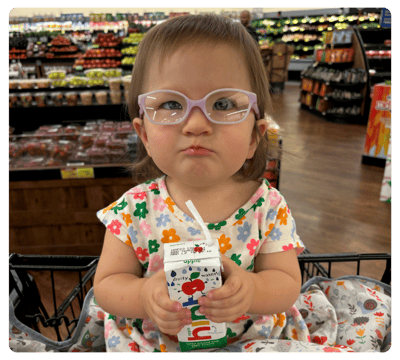 Young child wearing glasses sits in a shopping cart and holds a small water carton in a grocery store.