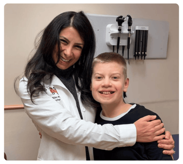 A clinician in a white coat hugs a smiling boy in a medical exam room.