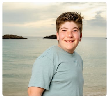 Smiling teenage boy in a light blue shirt standing beside the water.