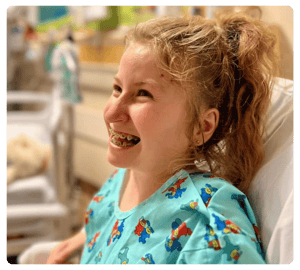 A smiling girl with braces sits in a hospital bed wearing a colorful gown.