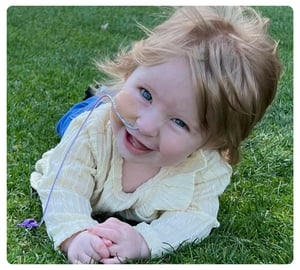 Smiling baby wearing a nasal feeding tube while lying on the grass.