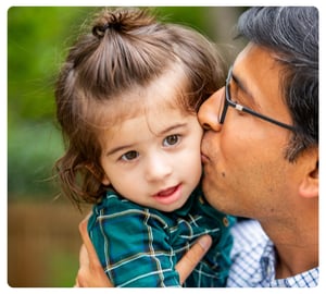 Father kissing his young child on the cheek while holding them outdoors.