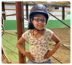 Young child wearing a riding helmet and glasses standing in an indoor horse arena.
