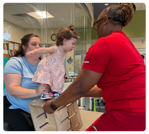 Toddler practicing standing during physical therapy with support from a caregiver and therapist.