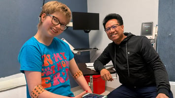 A smiling child wearing glasses and a bright blue T-shirt sits in a medical exam room beside a smiling healthcare provider. The child has patterned bandages on both arms, and medical equipment is visible in the background.