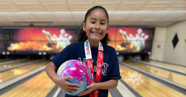A smiling young bowler stands in front of bowling lanes, holding a colorful pink and blue bowling ball. She wears a navy T-shirt and a Special Olympics medal around her neck.