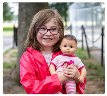 Young girl wearing glasses and a red jacket, smiling while holding a doll outdoors.