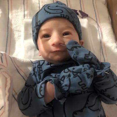 Newborn baby lying in a hospital bassinet, wearing a blue patterned outfit and matching cap with a feeding tube in their nose.