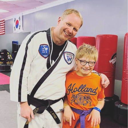 A smiling man in a white martial arts uniform stands with his arm around a smiling young boy wearing glasses, an orange T-shirt, and a purple belt. Red punching bags and flags are visible in the background of the martial arts studio.