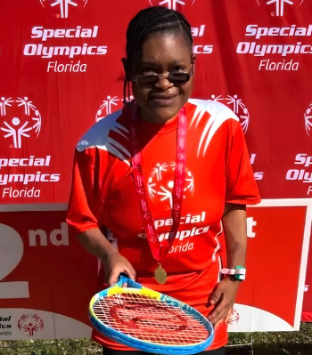 A smiling athlete wearing sunglasses, a red Special Olympics Florida shirt, and a gold medal holds a colorful tennis racket in front of a red Special Olympics backdrop.