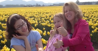 Three family members smiling together in a field of yellow flowers, with one child giving a thumbs-up and mountains in the background.