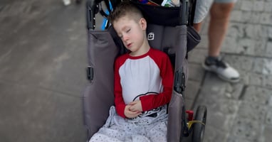 Child with Prader-Willi syndrome asleep in a stroller outdoors during the daytime.