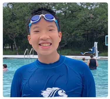 Young boy wearing swim goggles and a blue swim shirt, smiling at a swimming pool.