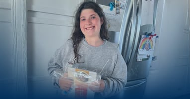 A smiling teenage girl with Prader-Willi syndrome stands in her kitchen holding a VYKAT XR package.