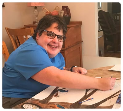 Smiling young man wearing glasses works on a drawing at a dining table.