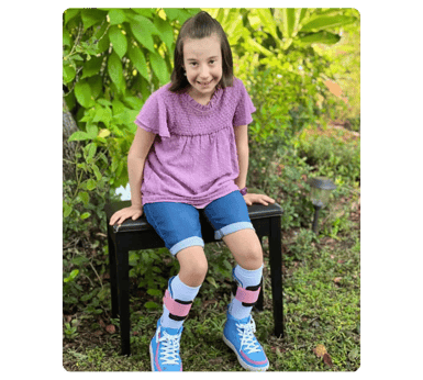 Young girl in a purple shirt and blue shorts sitting on a bench outdoors.