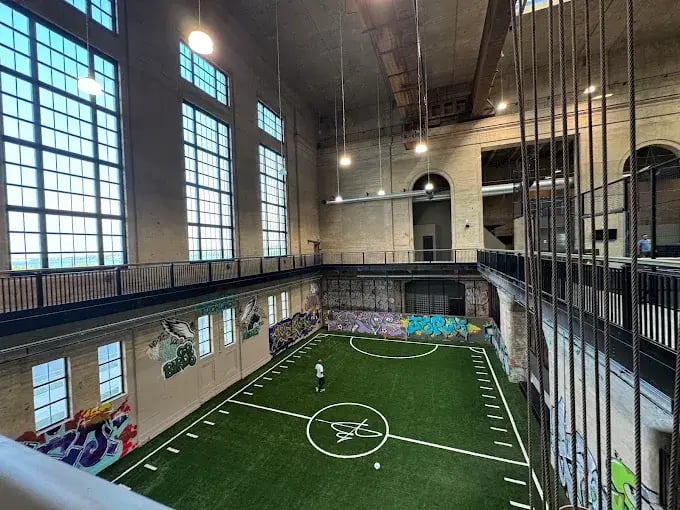 Indoor turf field at the event venue, viewed from an upper-level walkway with large windows and surrounding railings.
