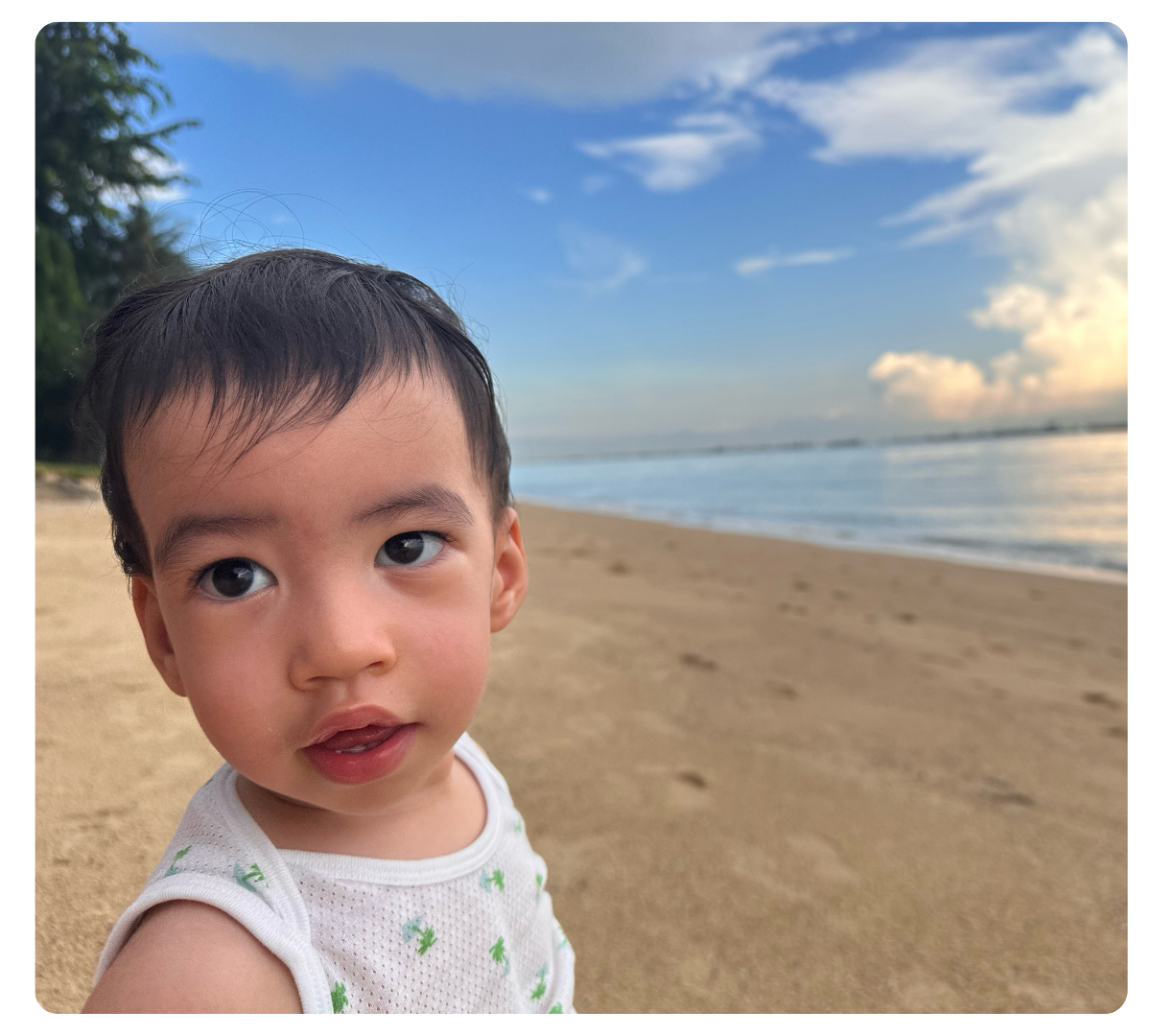 Young boy standing on a sandy beach with the ocean and blue sky behind him.