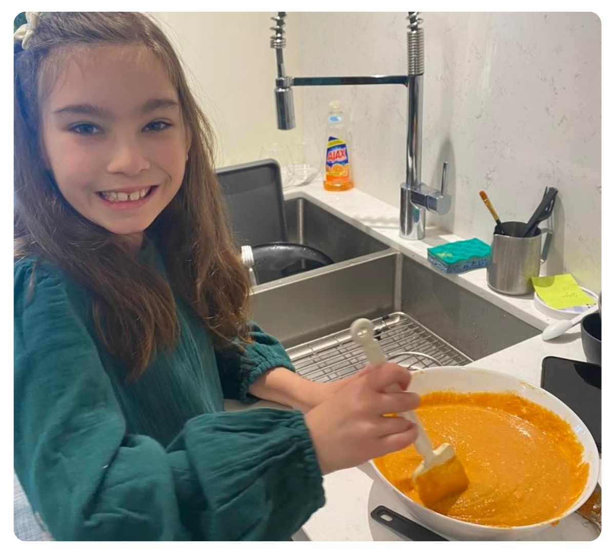 Young girl smiling while stirring orange batter in a mixing bowl at a kitchen sink.