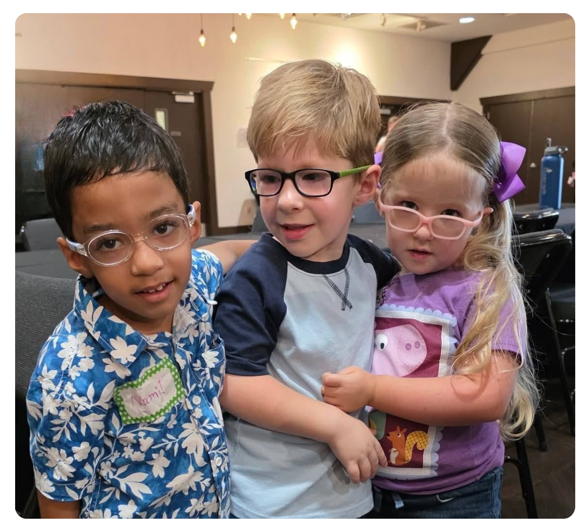 Three children wearing glasses standing close together and smiling at an indoor event.