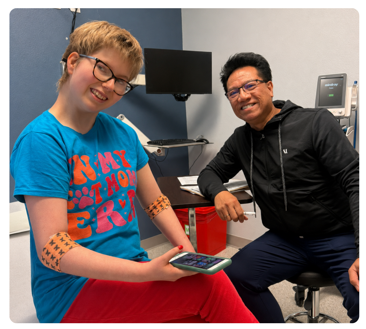 Young woman wearing glasses smiles beside a healthcare provider in an exam room, with monitoring patches on her arms.
