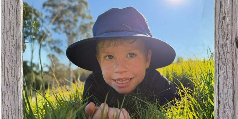 Young boy wearing a wide-brimmed hat lying in the grass and smiling at the camera.