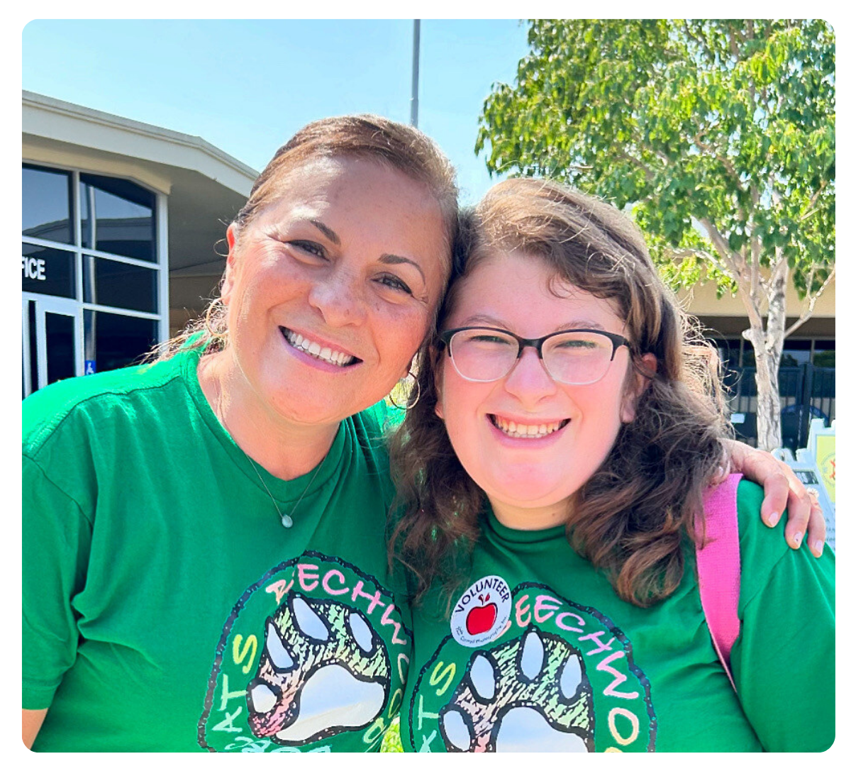 Two women in matching green shirts smile together outdoors, one wearing a volunteer badge.