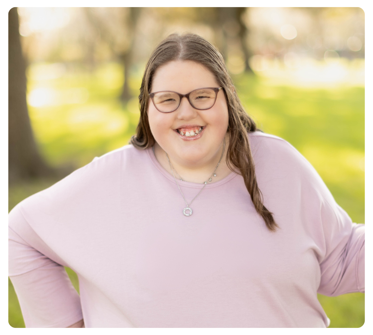 Young woman with glasses smiling outdoors in a light pink top.